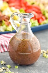 close up bottle of red wine vinaigrette with Cobb salad behind it and blue cheese and green onions around it