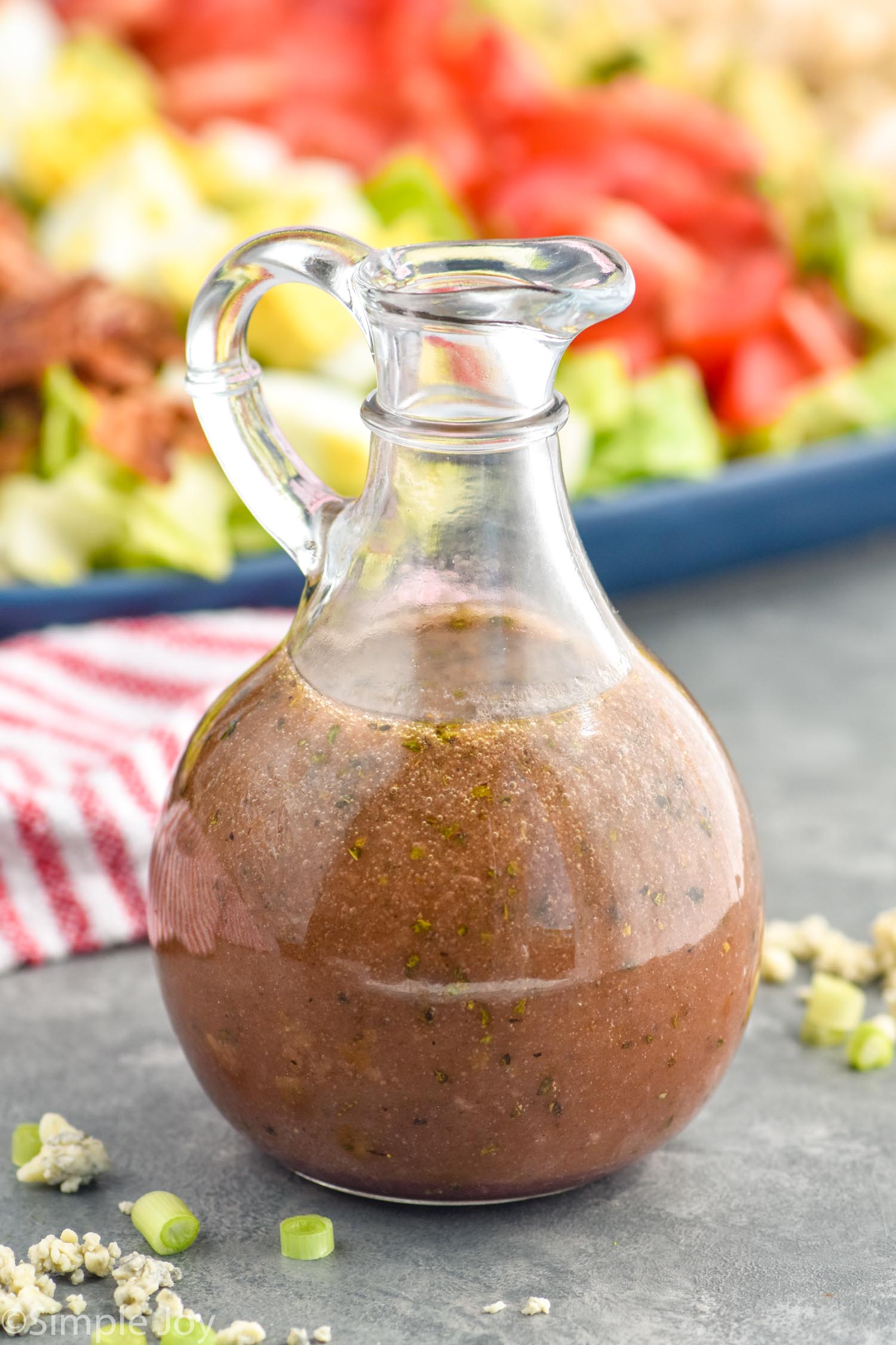 close up bottle of red wine vinaigrette with Cobb salad behind it and blue cheese and green onions around it