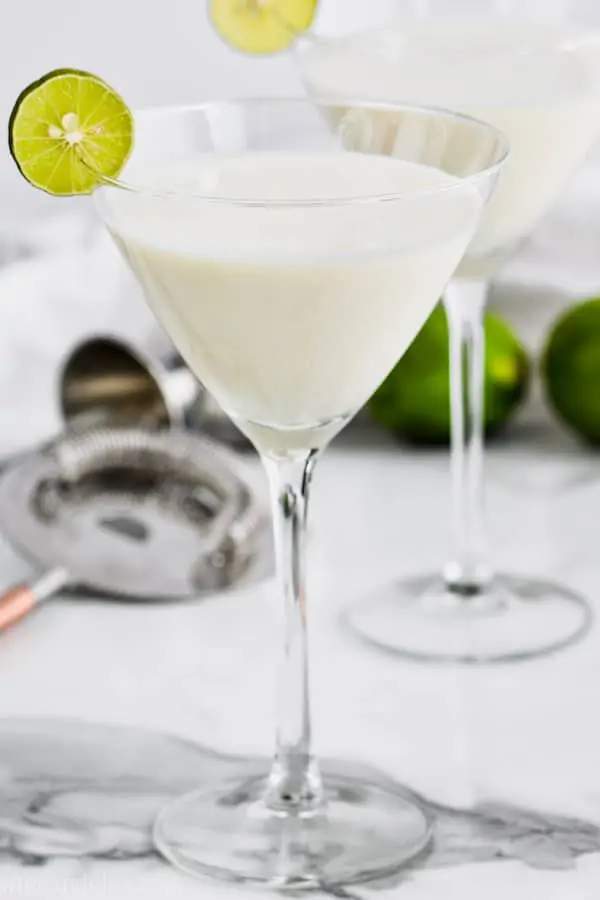 martini glass filled with white key lime martini recipe and garnished with a key lime slice, another glass, a cocktail strainer and limes in the background