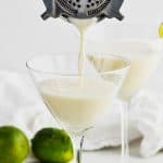 cocktail strainer pouring a white key lime martini recipe into a martini glass, another glass in the background that is full and garnished with a key lime wedge