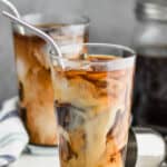 two tall tumblers with ice, iced coffee, and cream swirling in them with metal straws