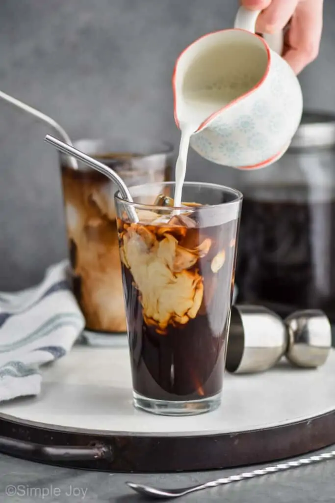 small cream pot pouring half and half into a large tumbler filled with ice and coffee to make an iced coffee