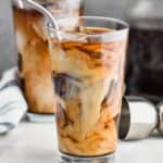 two tall tumblers with ice, iced coffee, and cream swirling in them with metal straws