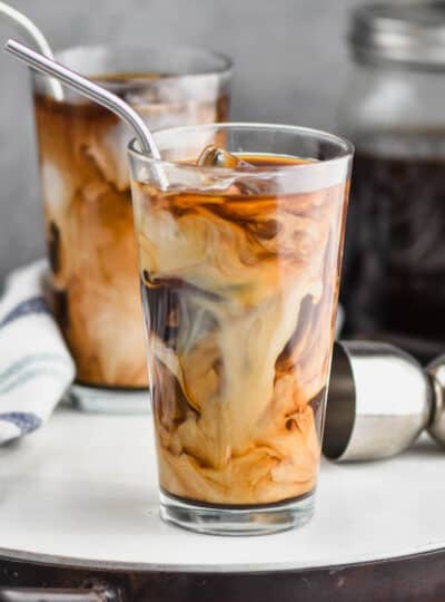 two tall tumblers with ice, iced coffee, and cream swirling in them with metal straws