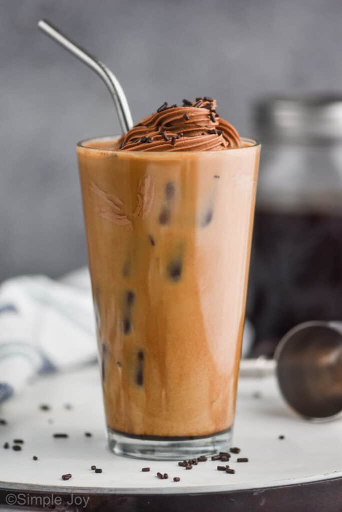 large glass tumbler completely full of ice and iced coffee topped with chocolate whipped cream and chocolate jimmies and a metal straw for an adult iced coffee cocktail