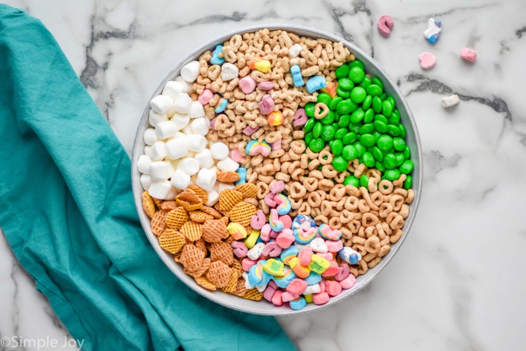 overhead of a bowl of ingredients broken up by ingredient, marshmallows, lucky charms, green m&ms, cheerios, lucky charms marshmallows, cirspix