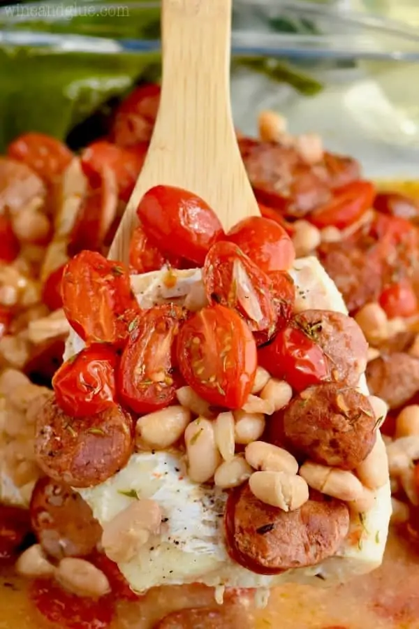 A wooden spoon is filled with roasted tomatoes, pinto beans, sausages, and fish. 