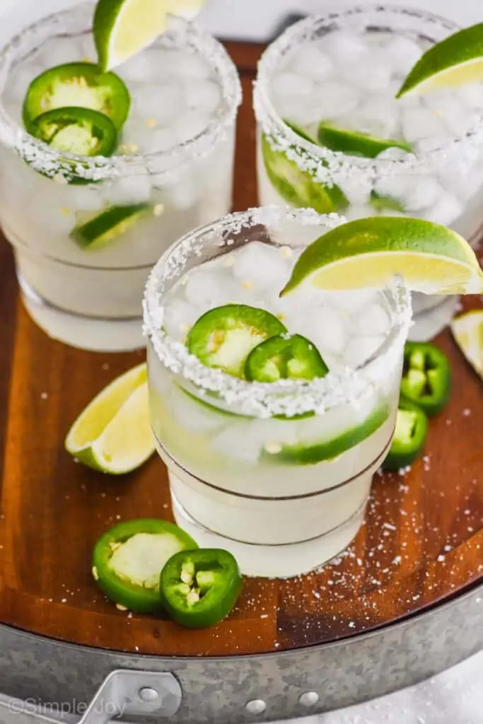 overhead of a wood tray with a metal rim that has three jalapeño margaritas in tumblers with salt rims, lime wedges, and fresh jalapeño slices