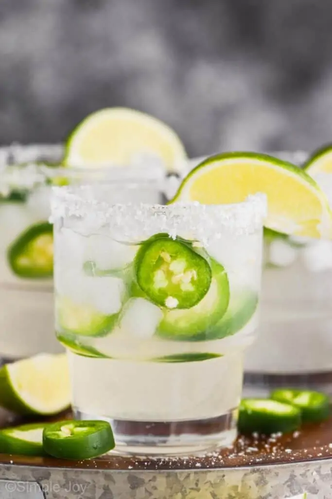 close up picture of a tumbler lined with salt, garnished with a lime wedge filled with a jalapeño margarita that has fresh jalapeño slices