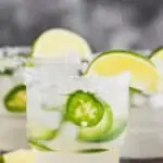 close up picture of a tumbler lined with salt, garnished with a lime wedge filled with a jalapeño margarita that has fresh jalapeño slices