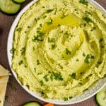 overhead view of a white bowl on a brown surface filled with edamame hummus recipe, garnished with parsley and oil and surrounded by cut vegetables and triangles of naan bread - 5