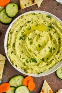 overhead view of a white bowl on a brown surface filled with edamame hummus recipe, garnished with parsley and oil and surrounded by cut vegetables and triangles of naan bread