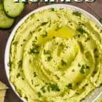 overhead view of a white bowl on a brown surface filled with edamame hummus recipe, garnished with parsley and oil and surrounded by cut vegetables and triangles of naan bread