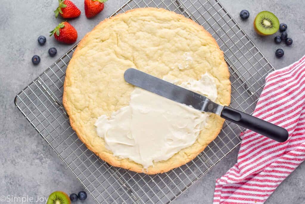 overhead view of a giant pizza sized sugar cookie that is being frosted with cream cheese frosting