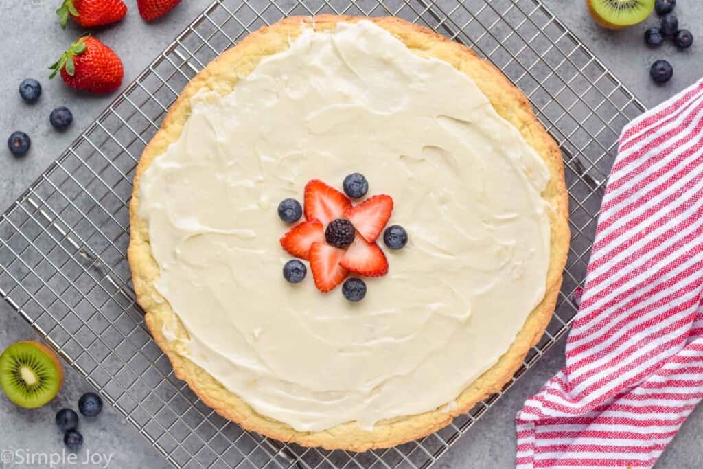 overhead view of a giant pizza sized sugar cookie that has been covered in cream cheese frosting and is starting to be decorated with fruit slices