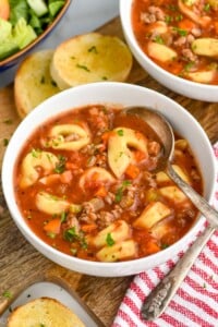 Overhead photo of Sausage Tortellini Soup served in a bowl with a spoon for eating. Slices of bread lay beside bowl.