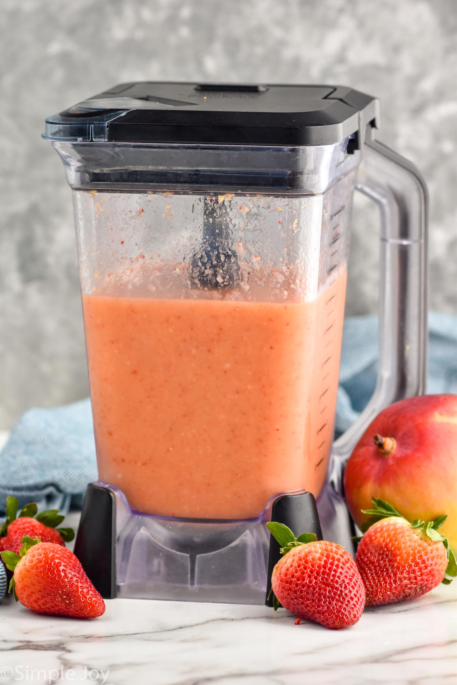 Photo of a blender of Strawberry Mango Margaritas recipe. Strawberries and a mango sit on counter beside blender.