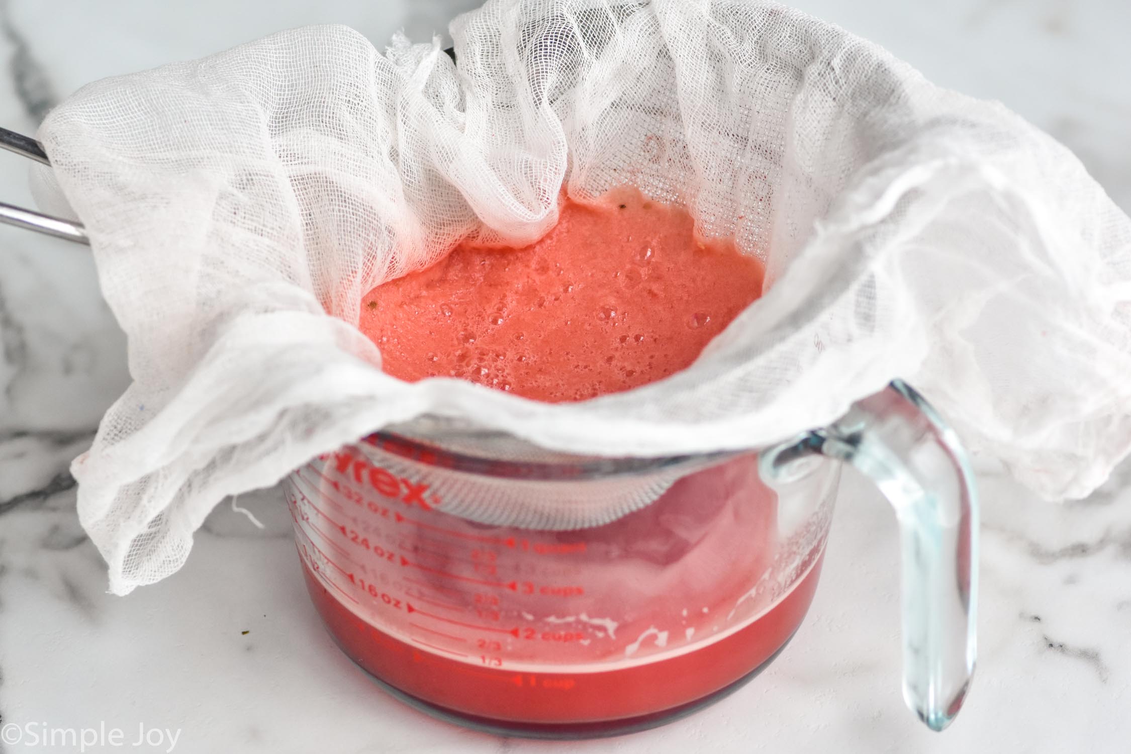 watermelon juice being strained through a wire mesh, lined with cheese cloth, into a measuring cup, view from into the cheesecloth