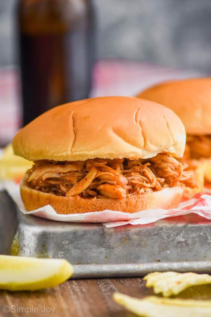 a bbq chicken sandwich served with potato chips, a pickle, and a beer in the background