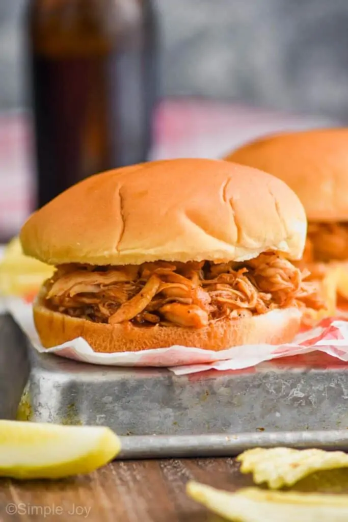 a bbq chicken sandwich served with potato chips, a pickle, and a beer in the background