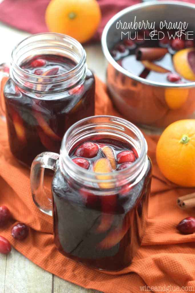 In mason jar glasses, the Cranberry Orange Mulled Wine has a dark maroon color with orange peels and cranberries. 