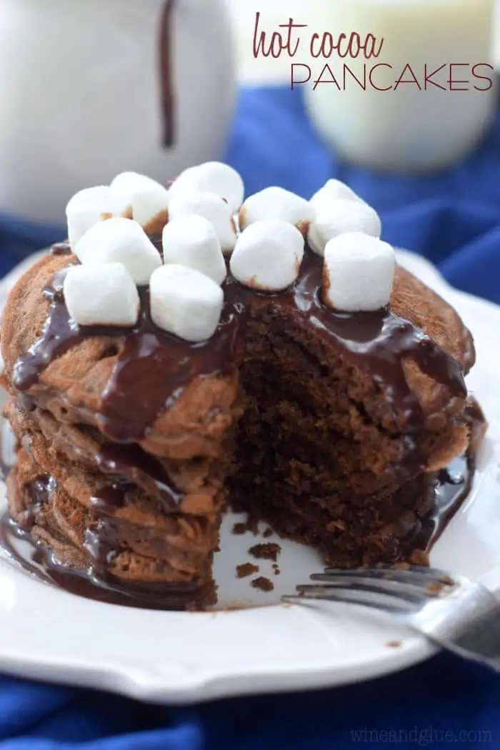 Stack of 5 Hot Cocoa Pancakes with chocolate drizzle and marshmallows on top. A small section in the shape of a triangle has been cut out. 