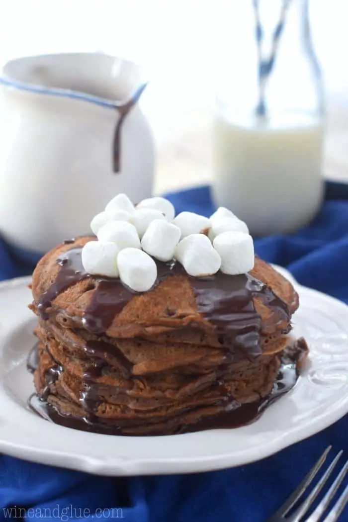 Stack of 5 Hot Cocoa Pancakes with chocolate drizzle and marshmallows on top. A small section in the shape of a triangle has been cut out. 