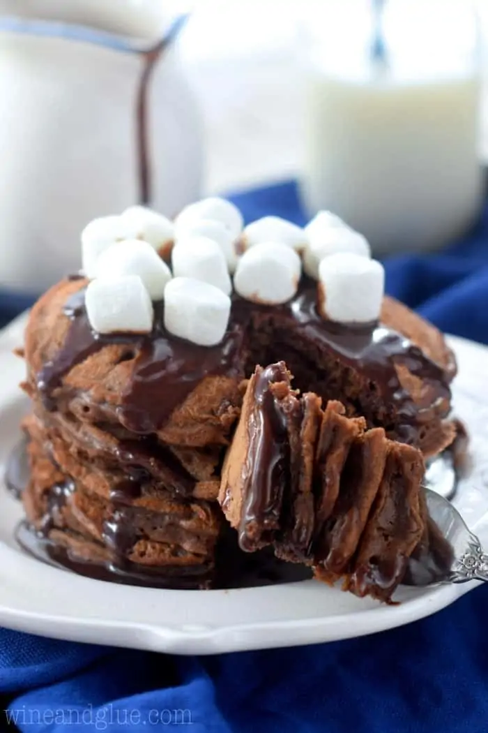 Stack of 5 Hot Cocoa Pancakes topped with chocolate drizzle and marshmallows on top while a fork is taking a section. 