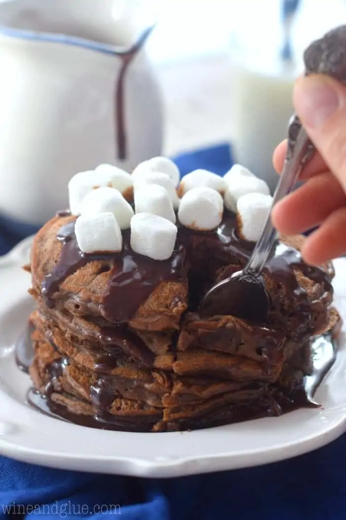 Stack of 5 Hot Cocoa Pancakes topped with chocolate drizzle and marshmallows on top while a fork is taking a section. 