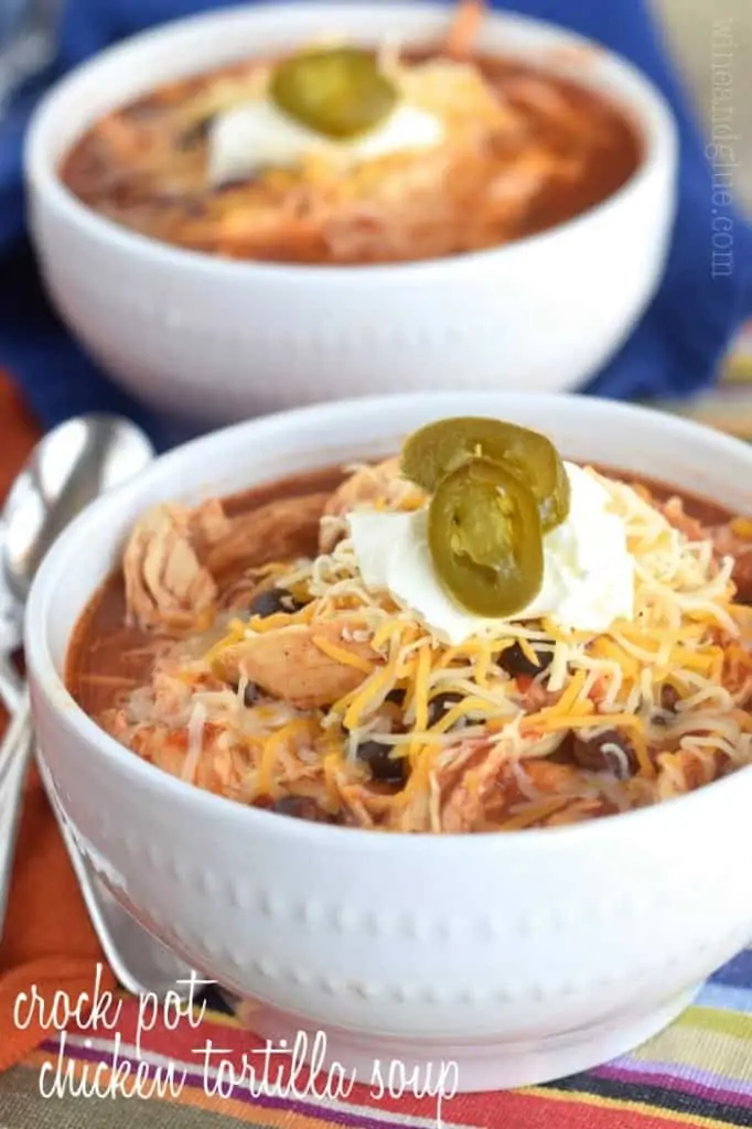 In a white bowl, the Crockpot Taco Soup is topped with shredded cheese, sour cream, and pickled jalapenos. 