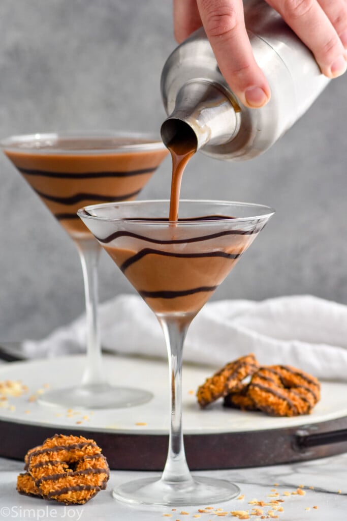 Photo of person's hand pouring Samoa Martini recipe into martini glass garnished with chocolate. Samoa cookies on counter beside martini glasses.