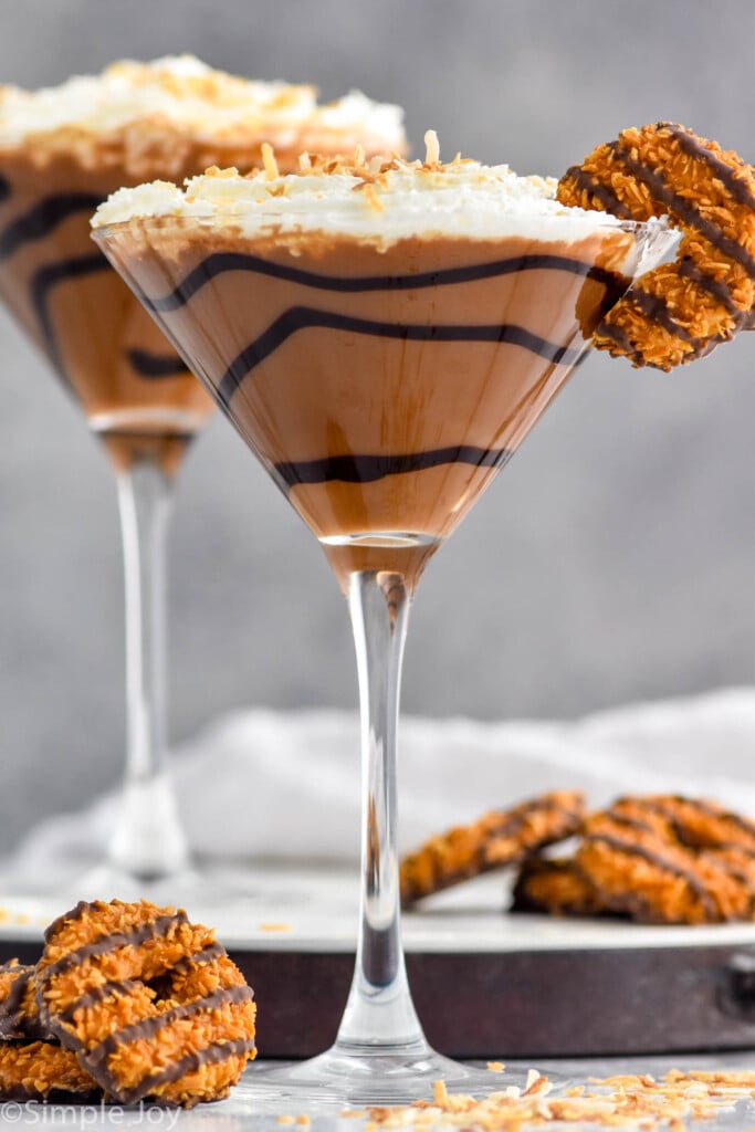 Photo of two Samoa Martinis garnished with Samoa cookies. Extra Samoa cookies on the counter beside Samoa Martinis.