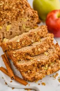 close up of cinnamon apple bread with streusel bread that has been sliced into on a marble cutting board with cinnamon sticks next to it and apples in the background