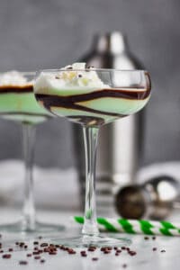a tall coup glass lined with chocolate syrup filled with a green grasshopper cocktail and chocolate sprinkles around the base of the glass