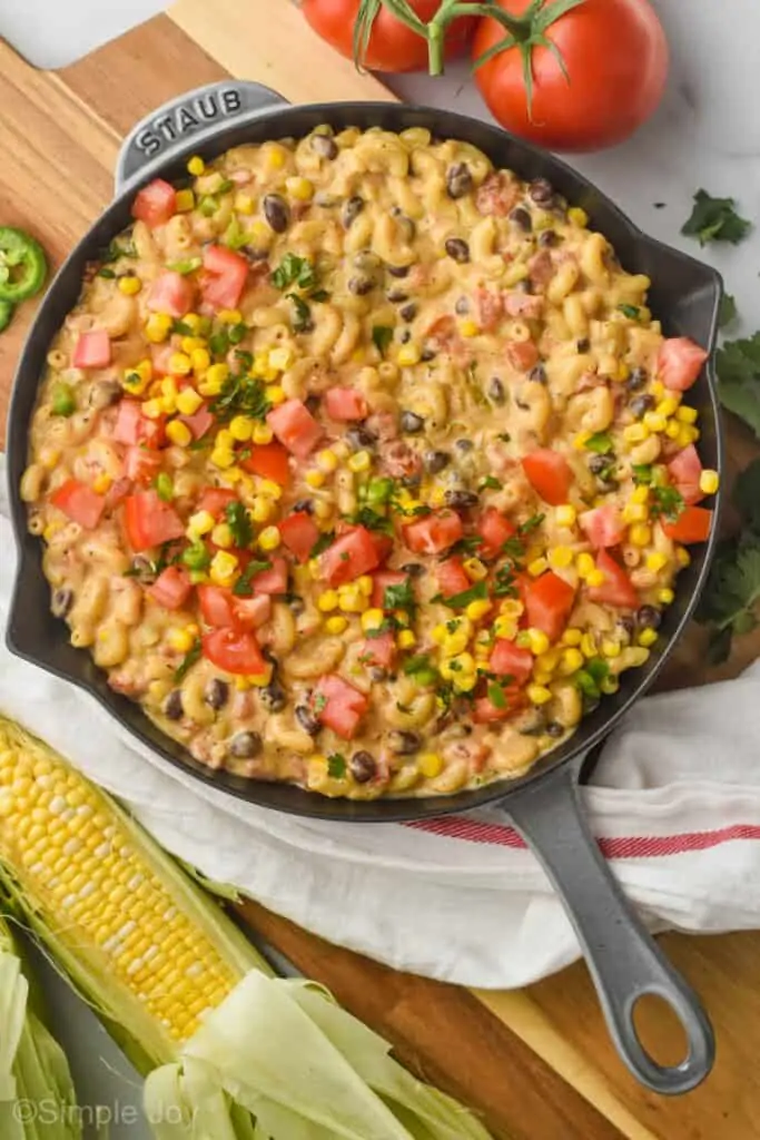 overhead view of a gray skillet filled with southwestern macaroni and cheese recipe topped with fresh tomatoes, corn, and jalapeños on a cutting board surrounded by fresh ingredients