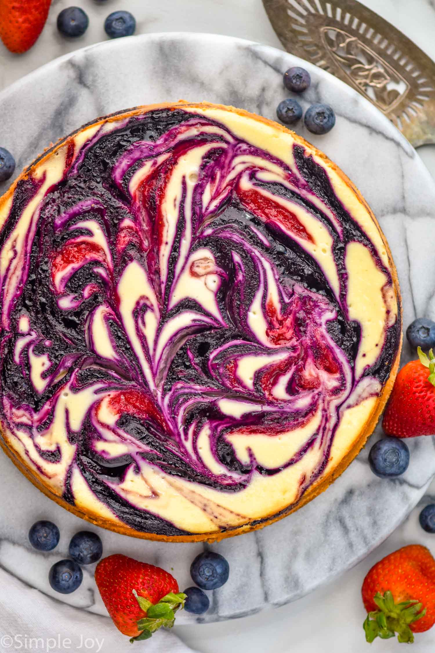 Overhead photo of Berry Cheesecake on a platter with blueberries and strawberries. Serving spatula next to platter.