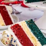 close up of a slice of red white and blue cake on three stacked white plates with the rest of the cake in the background