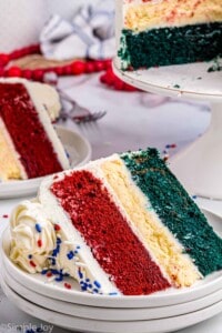 close up of a slice of red white and blue cake on three stacked white plates with the rest of the cake in the background