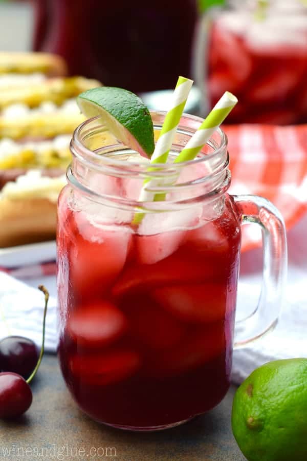 In a mason jar glass, the Cherry Limeade has a dark red color with ice cubes and a slice of lime on the rim. 