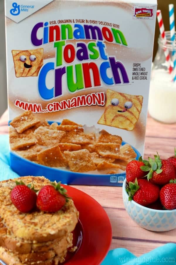 The stack of Cinnamon Toast Crunch Crusted French Toast with two strawberries in Front. In the background, a box of Cinnamon Toast Crunch is shown with a bowl of strawberries. 