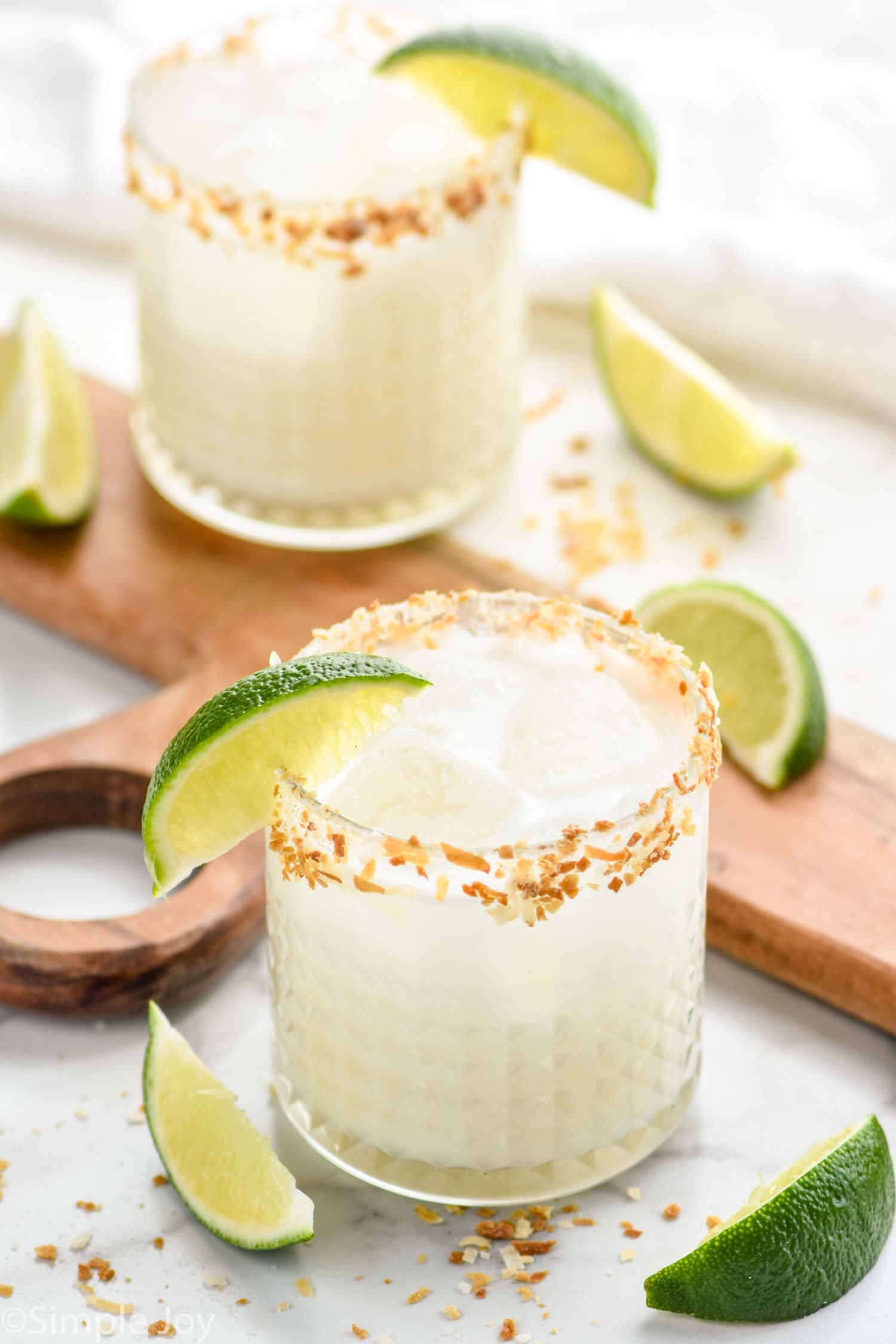two coconut margaritas on a table surrounded with lime wedges and toasted coconut
