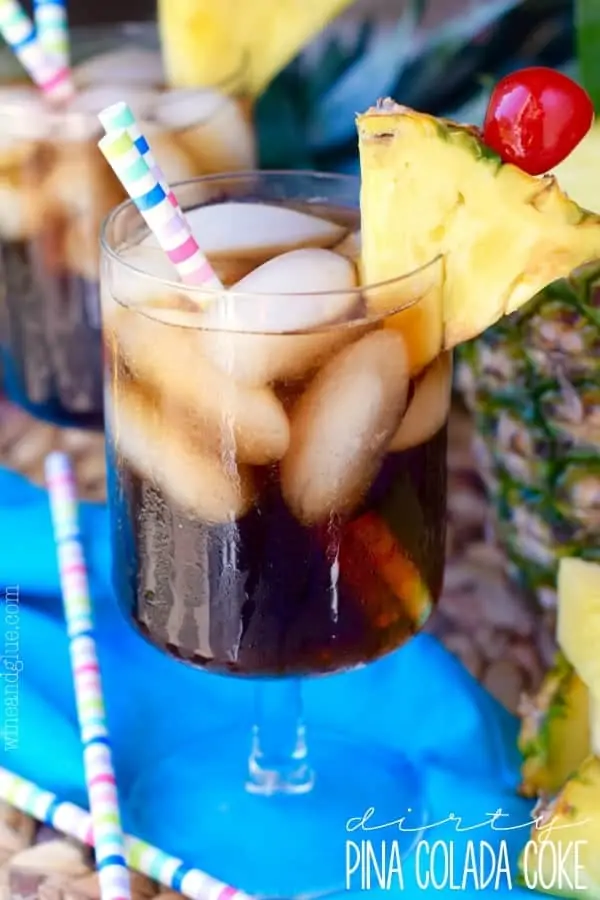 In a glass, the Dirty Pina Colada Coke has two paper straws and rimed with a sliced pinapple and cherry. 