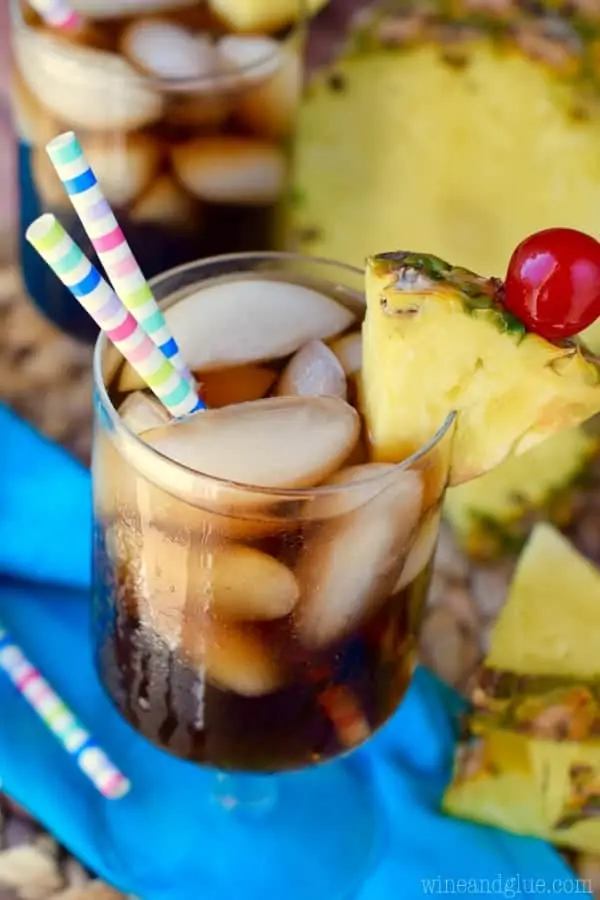 In a glass, the Dirty Pina Colada Coke has two paper straws and rimed with a sliced pinapple and cherry. 