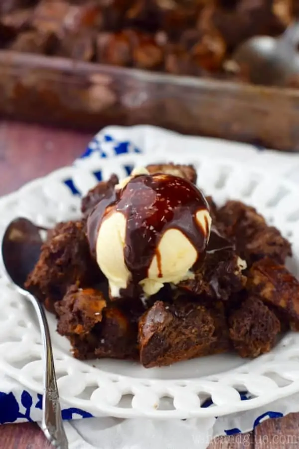 On a white plate, the Chocolate Pretzel Bread Pudding has a scoop of vanilla ice cream and chocolate drizzle with flaky salt sprinkled on top. 