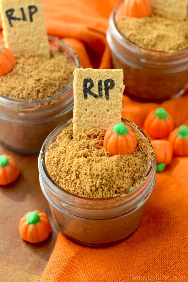 In a small mason jar, the Pumpkin spice Chocolate Pudding is topped with crushed graham cracker, a pumpkin candy, and a stick of a graham cracker with the letter RIP. 