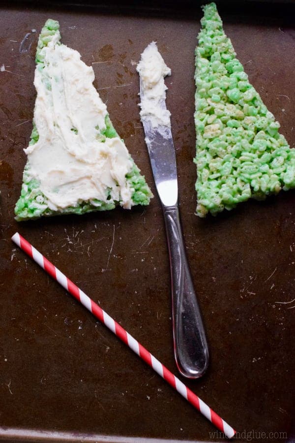Two triangular green Rice Krispies are side to side. One of them has white frosting spread on top of it. 