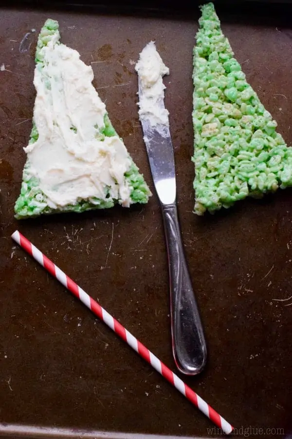 Two triangular green Rice Krispies are side to side. One of them has white frosting spread on top of it. 