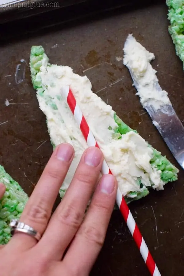 On a triangular green Rice Krispies, white frosting is spread on top and a woman is placing a paper straw with red strips in the middle of the frosted Rice Krispie.. 