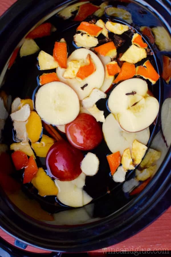 An overhead photo of a Slow Cooker with wine inside and cuts of apples, orange peels, lemon peels, and cinnamon. 