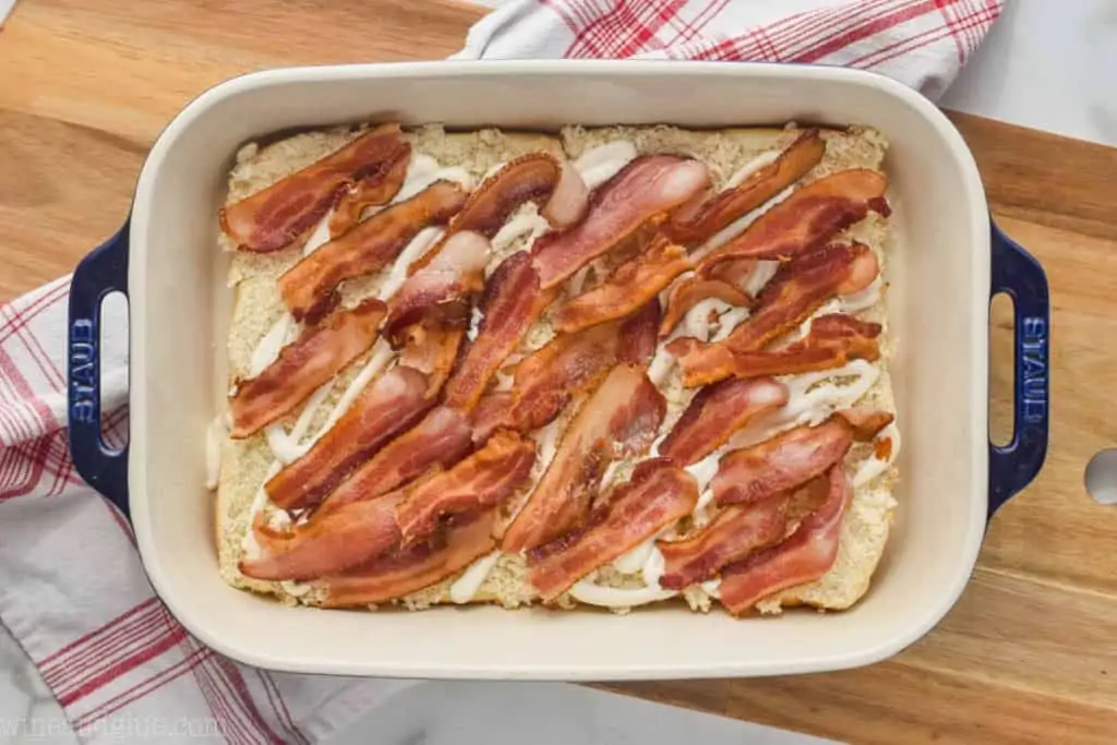 landscape photo of a baking dish on a cutting board with the bottom half of slider rolls, topped with cooked bacon - 3